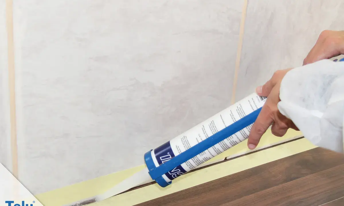 Eine Hand zieht mit einer Kartusche präzise silikonfugen. Klebeband schützt den Boden und die Wandfliesen.