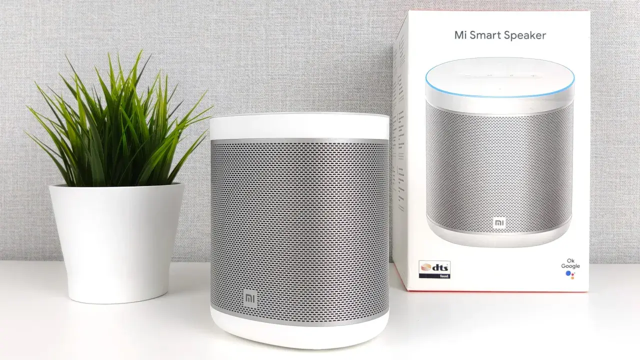Ein weißer Mi Smart Speaker steht neben einer grünen Pflanze. Die Verpackung des Lautsprechers ist ebenfalls zu sehen.