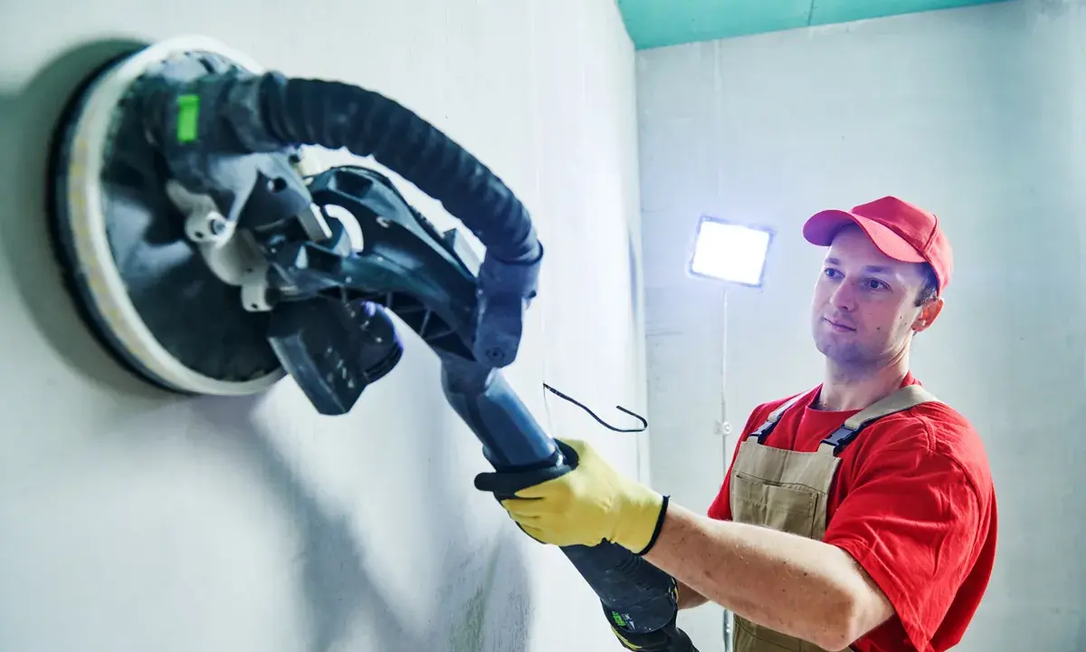 Mann schleift mit einem Langhalsschleifer die Wand per Hand.