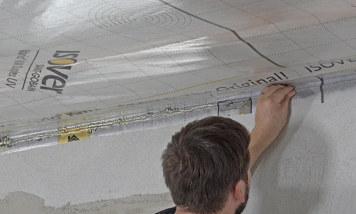 Mann bringt eine Dampfbremse an der Decke an. Die silberne Folie mit dem Isover-Logo wird sorgfältig verklebt.