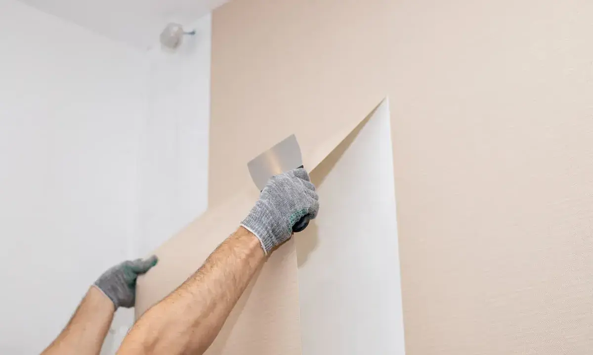 Hände mit Handschuhen ziehen mit einem Spachtel die alte Vliestapete ab.