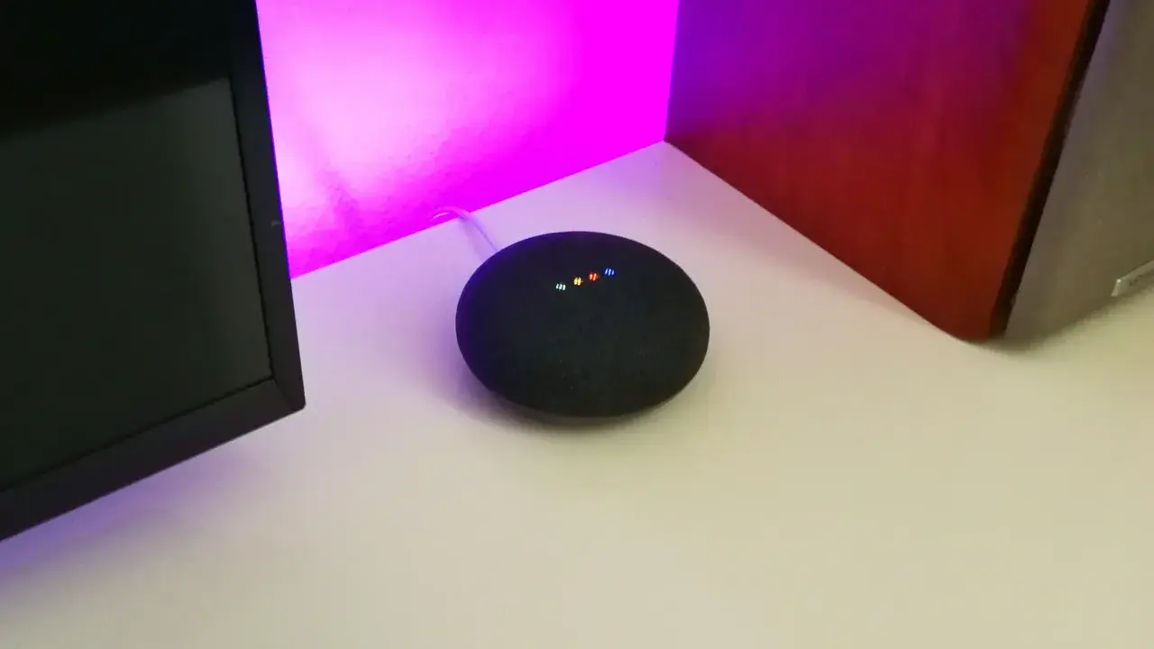 Ein Google Nest Mini mit leuchtenden Punkten, bereit für die Einrichtung. Der smarte Lautsprecher steht auf einem Schreibtisch neben einem Monitor und einer roten Wand.