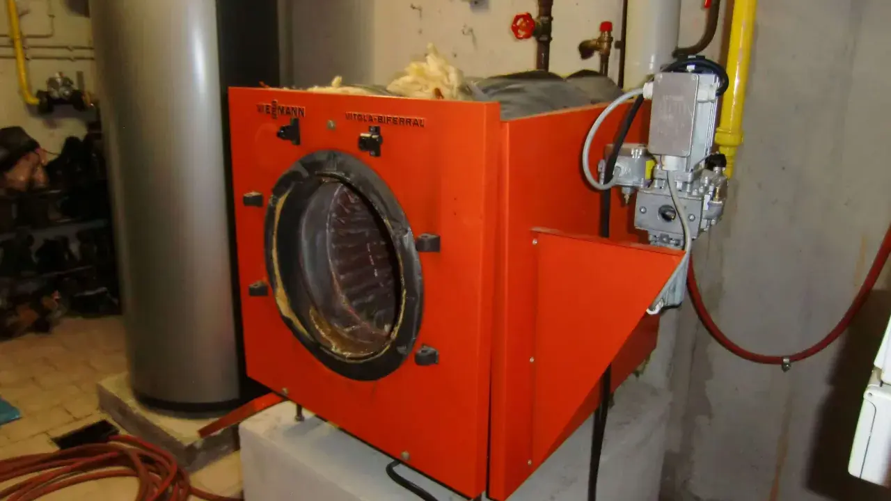 Alte Gasheizung in orange, daneben ein grauer Warmwasserspeicher. Rohre und ein Druckmesser sind sichtbar.