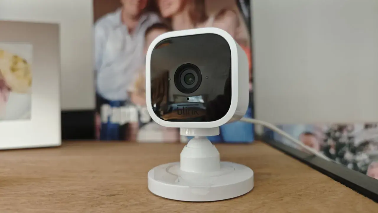 Kompakte Überwachungskamera mit Blink-Logo, die als Alexa-kompatible Alarmanlage dient. Im Hintergrund sind Familienfotos zu sehen.