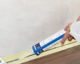 Eine Hand zieht mit einer Kartusche präzise silikonfugen. Klebeband schützt den Boden und die Wandfliesen.