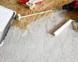 Beim Streichen von OSB-Platten mit weißer Farbe: Ein Farbroller und eine Farbwanne liegen bereit, daneben ein Akkuschrauber und Handschuhe.