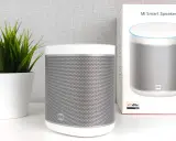 Ein weißer Mi Smart Speaker steht neben einer grünen Pflanze. Die Verpackung des Lautsprechers ist ebenfalls zu sehen.