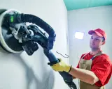 Mann schleift mit einem Langhalsschleifer die Wand per Hand.