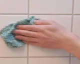 Hand wischt mit einem Tuch über weiße Wandfliesen, um sie zu reinigen.