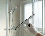 Mit einem Gummiabzieher wird die nasse Glasdusche gereinigt. Wasser und Schaum laufen über das Glas.