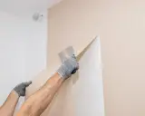 Hände mit Handschuhen ziehen mit einem Spachtel die alte Vliestapete ab.