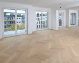 Ein heller Fischgrätenparkettboden, frisch nach dem Parkett schleifen, erstrahlt in einem modernen Wohnraum mit großen Fenstern und Balkonen.