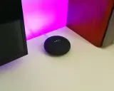 Ein Google Nest Mini mit leuchtenden Punkten, bereit für die Einrichtung. Der smarte Lautsprecher steht auf einem Schreibtisch neben einem Monitor und einer roten Wand.