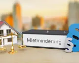 Hausmodell, Waage und Ordner mit "Mietminderung" Schriftzug. Ein Paragrafensymbol deutet auf rechtliche Aspekte hin, z.B. bei Problemen mit der Heizung.