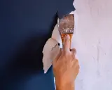 Hand schabt mit einem Spachtel die alte, weiße Dispersionsfarbe von einer dunkelblauen Wand.