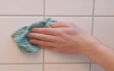 Hand wischt mit einem Tuch über weiße Wandfliesen, um sie zu reinigen.