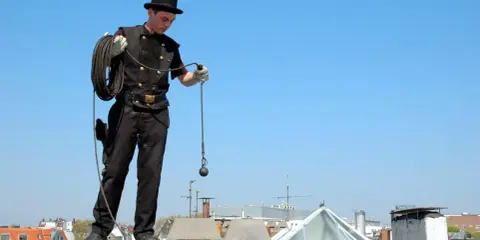 Ein Schornsteinfeger in traditioneller Kleidung mit Zylinder und Seil steht auf einem Dach. Die Reinigung der Kamine ist eine wichtige Schornsteinfeger Pflicht.