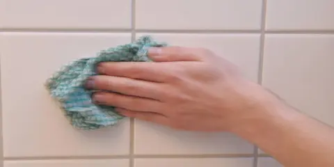Hand wischt mit einem Tuch über weiße Wandfliesen, um sie zu reinigen.