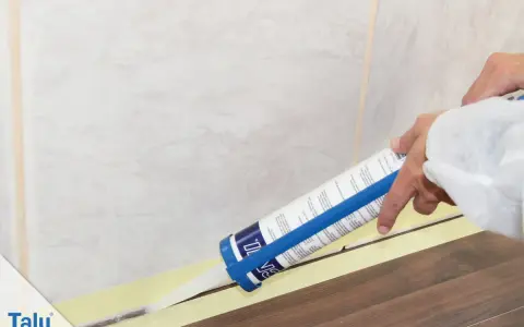 Eine Hand zieht mit einer Kartusche präzise silikonfugen. Klebeband schützt den Boden und die Wandfliesen.
