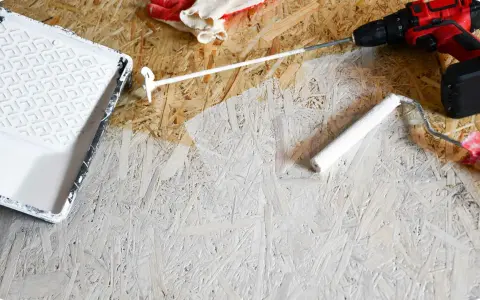 Beim Streichen von OSB-Platten mit weißer Farbe: Ein Farbroller und eine Farbwanne liegen bereit, daneben ein Akkuschrauber und Handschuhe.