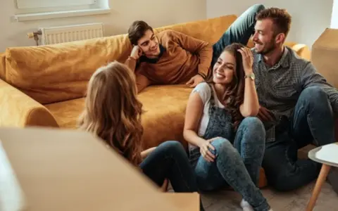 Glückliche Freunde lachen auf dem Boden vor einem Sofa, umgeben von Umzugskartons. Ein privater Vermieter könnte ihnen hier beim Einzug helfen.