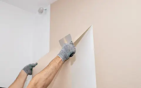 Hände mit Handschuhen ziehen mit einem Spachtel die alte Vliestapete ab.