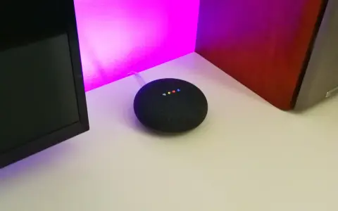 Ein Google Nest Mini mit leuchtenden Punkten, bereit für die Einrichtung. Der smarte Lautsprecher steht auf einem Schreibtisch neben einem Monitor und einer roten Wand.