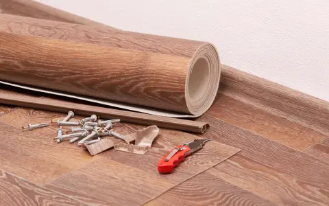 Materialien für das Linoleum verlegen: eine gerollte Bodenbelagsbahn, Sockelleisten, Dübel, Schrauben und ein Cuttermesser liegen bereit.