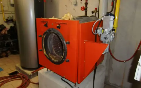 Alte Gasheizung in orange, daneben ein grauer Warmwasserspeicher. Rohre und ein Druckmesser sind sichtbar.