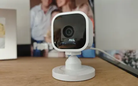 Kompakte Überwachungskamera mit Blink-Logo, die als Alexa-kompatible Alarmanlage dient. Im Hintergrund sind Familienfotos zu sehen.
