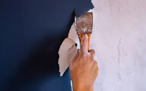 Hand schabt mit einem Spachtel die alte, weiße Dispersionsfarbe von einer dunkelblauen Wand.