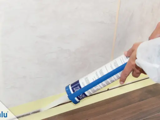 Eine Hand zieht mit einer Kartusche präzise silikonfugen. Klebeband schützt den Boden und die Wandfliesen.
