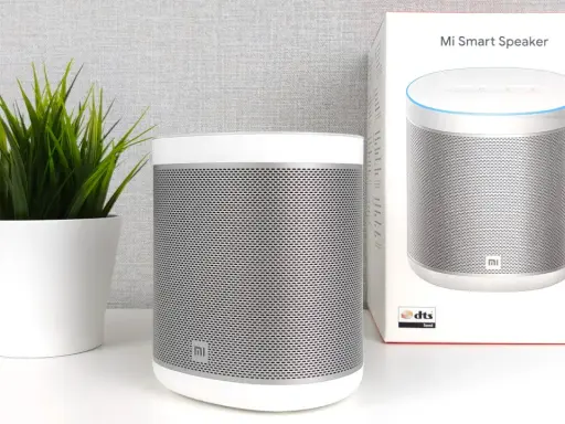 Ein weißer Mi Smart Speaker steht neben einer grünen Pflanze. Die Verpackung des Lautsprechers ist ebenfalls zu sehen.