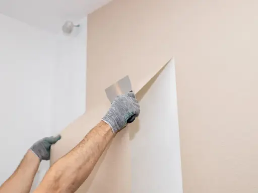 Hände mit Handschuhen ziehen mit einem Spachtel die alte Vliestapete ab.
