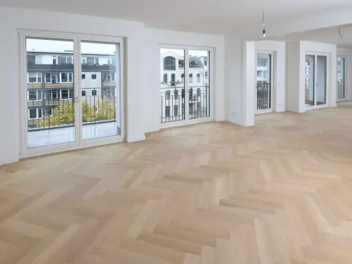 Ein heller Fischgrätenparkettboden, frisch nach dem Parkett schleifen, erstrahlt in einem modernen Wohnraum mit großen Fenstern und Balkonen.