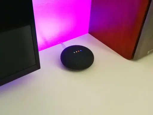 Ein Google Nest Mini mit leuchtenden Punkten, bereit für die Einrichtung. Der smarte Lautsprecher steht auf einem Schreibtisch neben einem Monitor und einer roten Wand.