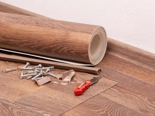 Materialien für das Linoleum verlegen: eine gerollte Bodenbelagsbahn, Sockelleisten, Dübel, Schrauben und ein Cuttermesser liegen bereit.