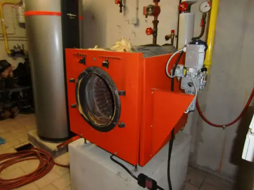 Alte Gasheizung in orange, daneben ein grauer Warmwasserspeicher. Rohre und ein Druckmesser sind sichtbar.