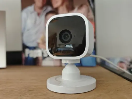 Kompakte Überwachungskamera mit Blink-Logo, die als Alexa-kompatible Alarmanlage dient. Im Hintergrund sind Familienfotos zu sehen.