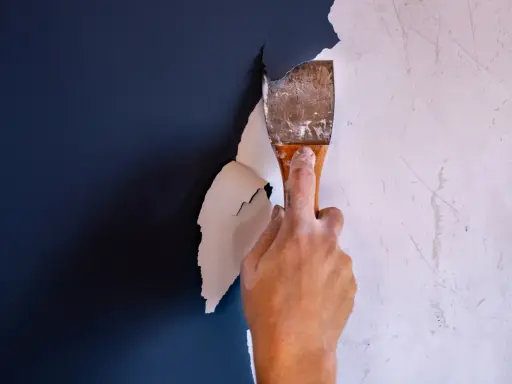 Hand schabt mit einem Spachtel die alte, weiße Dispersionsfarbe von einer dunkelblauen Wand.