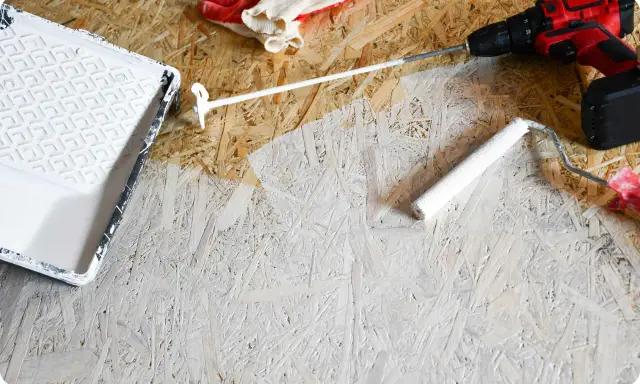 Beim Streichen von OSB-Platten mit weißer Farbe: Ein Farbroller und eine Farbwanne liegen bereit, daneben ein Akkuschrauber und Handschuhe.