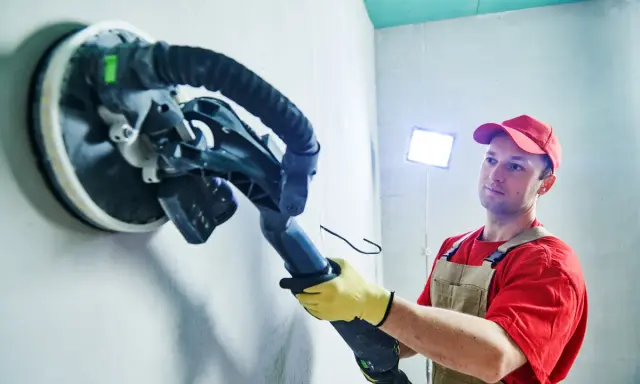 Mann schleift mit einem Langhalsschleifer die Wand per Hand.