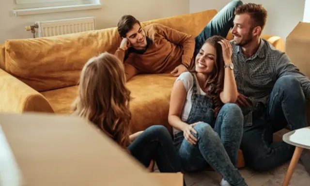 Glückliche Freunde lachen auf dem Boden vor einem Sofa, umgeben von Umzugskartons. Ein privater Vermieter könnte ihnen hier beim Einzug helfen.