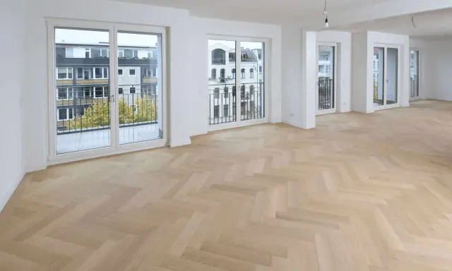 Ein heller Fischgrätenparkettboden, frisch nach dem Parkett schleifen, erstrahlt in einem modernen Wohnraum mit großen Fenstern und Balkonen.