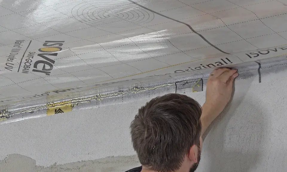 Mann bringt eine Dampfbremse an der Decke an. Die silberne Folie mit dem Isover-Logo wird sorgfältig verklebt.