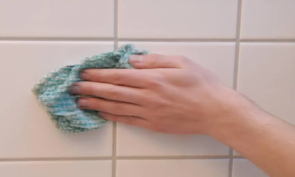 Hand wischt mit einem Tuch über weiße Wandfliesen, um sie zu reinigen.