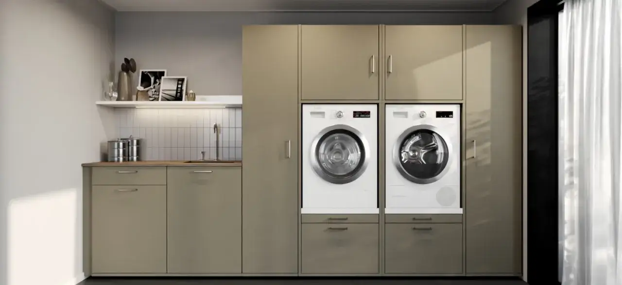 Moderne Waschküche mit Waschmaschine und trockner nebeneinander, eingebaut in beige Schränke. Links eine Spüle mit Arbeitsplatte.