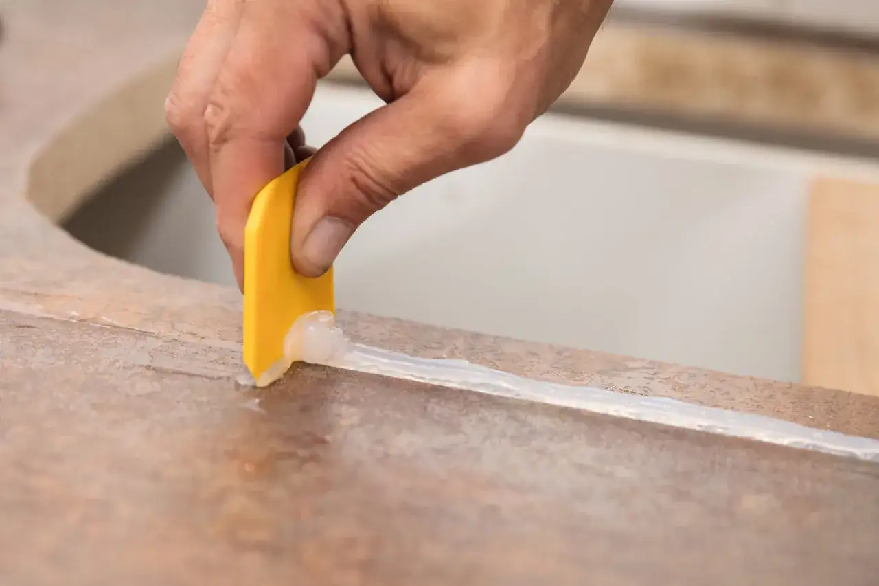 Hand zieht mit gelbem Werkzeug eine Silikonfuge auf einer Arbeitsplatte.