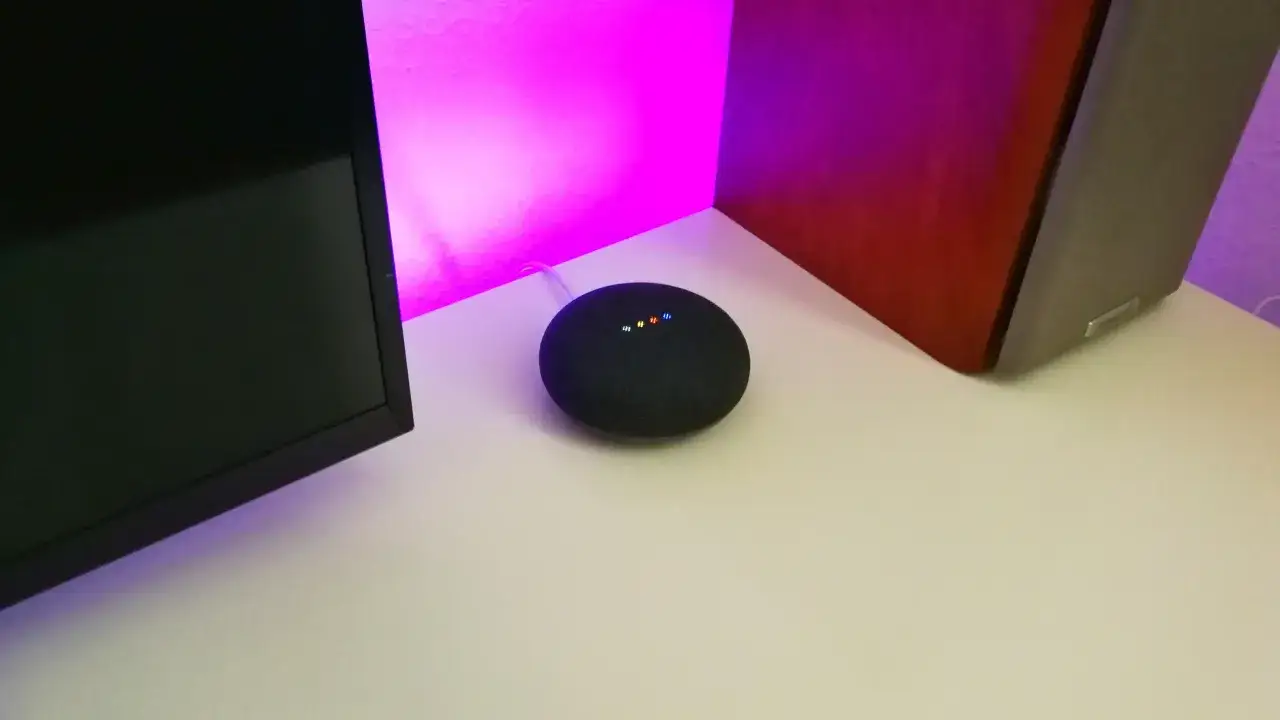 Ein schwarzer Google Nest Mini mit leuchtenden Punkten, bereit für die Einrichtung, steht neben einem Monitor und einem Lautsprecher.