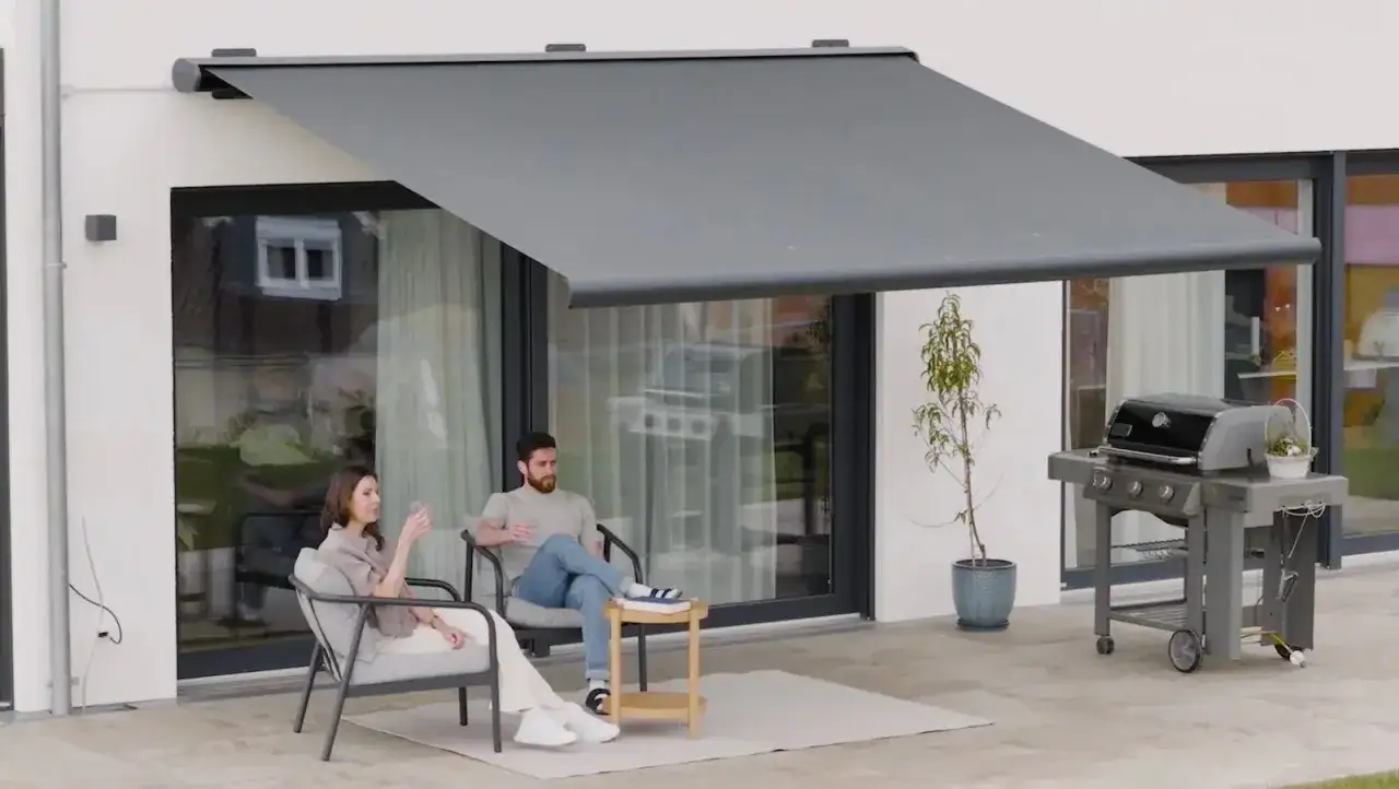 Ein Paar entspannt sich unter einer modernen Markise auf einer Terrasse. Ein Grill steht bereit für ein Sommerfest.